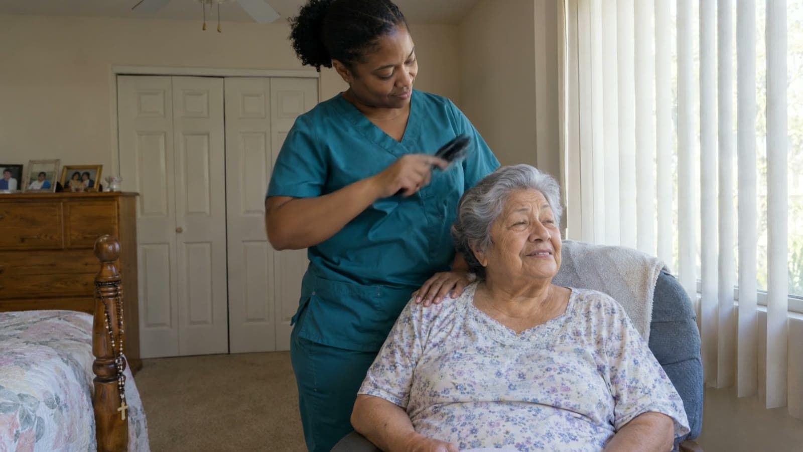 An aide in teal scrubs brushes the gray hair of an older woman sitting in a chair by a bedroom window. The older woman is smiling.